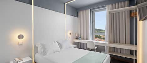 A modern hotel room with a bed, bedside table with a phone, a desk with a chair, a window with curtains, and a built-in wardrobe.