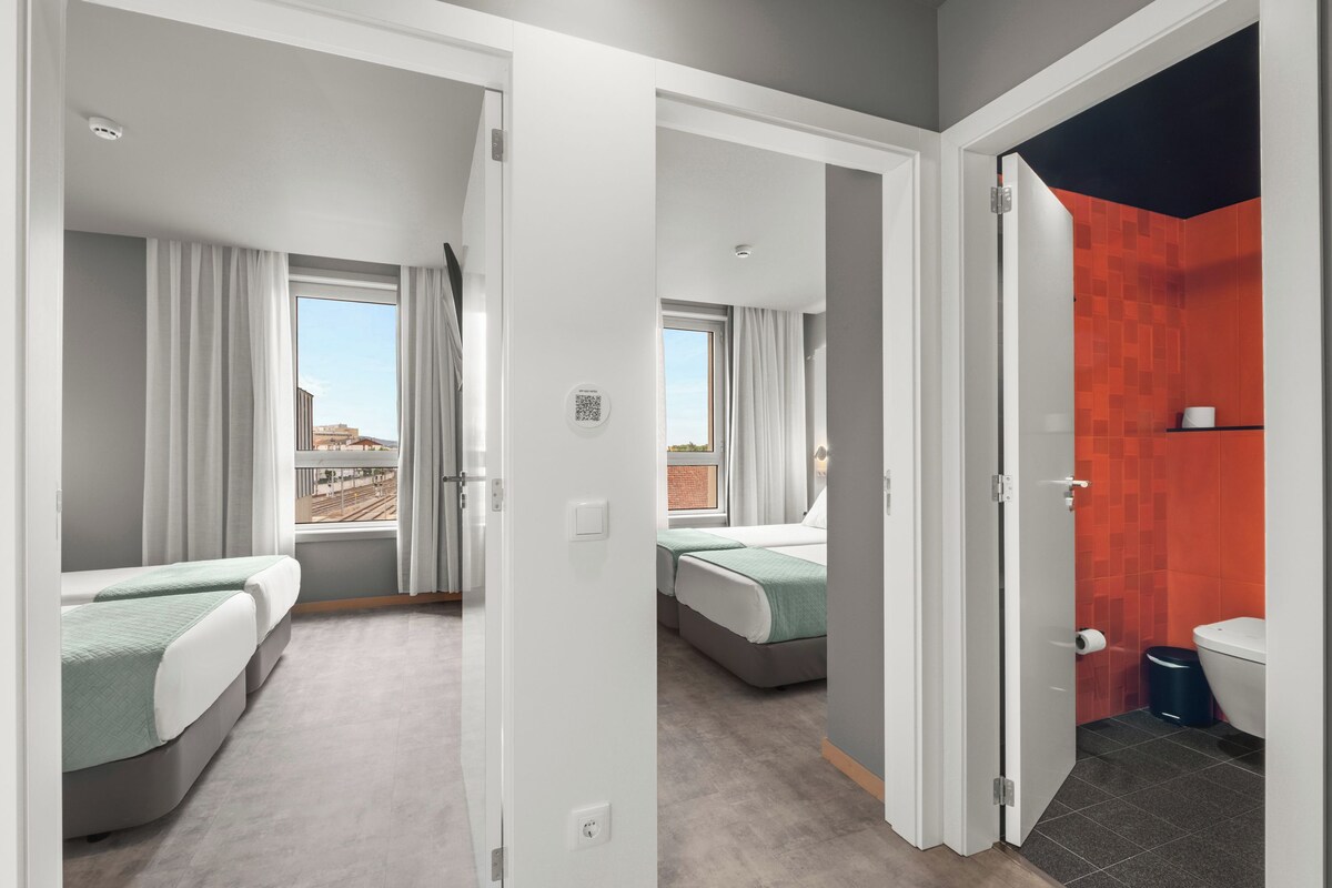 A modern hotel room with two beds, a bathroom with red tiles, and a view of the cityscape through the windows.