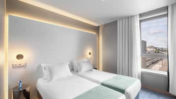 Habitación de hotel con dos camas, una mesita de noche y una ventana con cortinas, ofreciendo vistas al exterior.