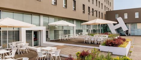 Une terrasse extérieure avec des tables et des chaises blanches, entourée d’une végétation luxuriante et bordée d’une architecture moderne.