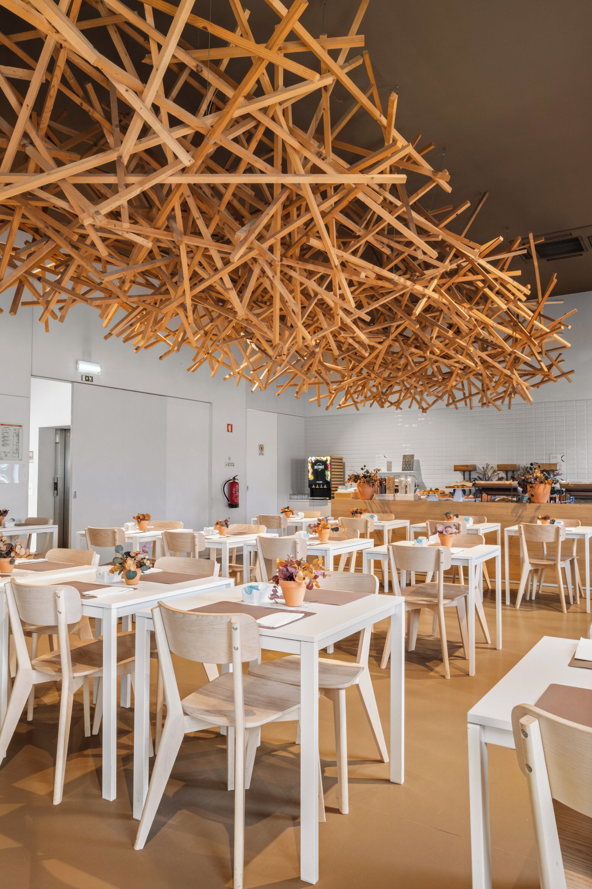 A modern dining area with white tables and chairs, a wooden ceiling structure, and a kitchen in the background.