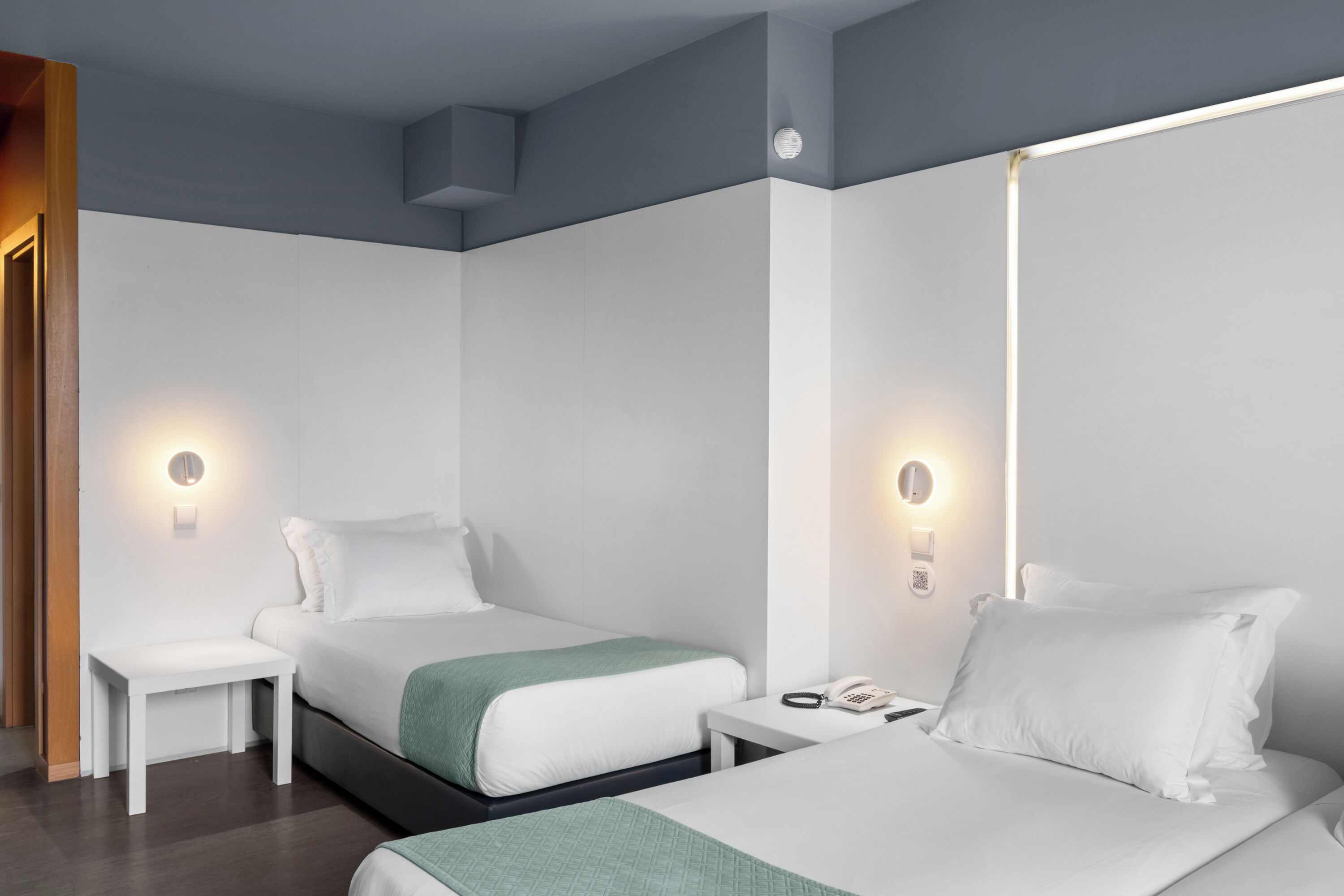 A modern hotel room with two beds, a bedside table with a phone, and wall-mounted light fixtures.