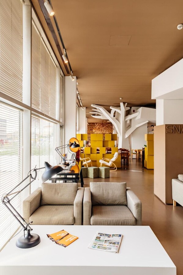 A modern interior with a white desk, a lamp, and a magazine. There are two beige sofas and a yellow bookshelf in the background. - Basic Braga by Axis (Braga)