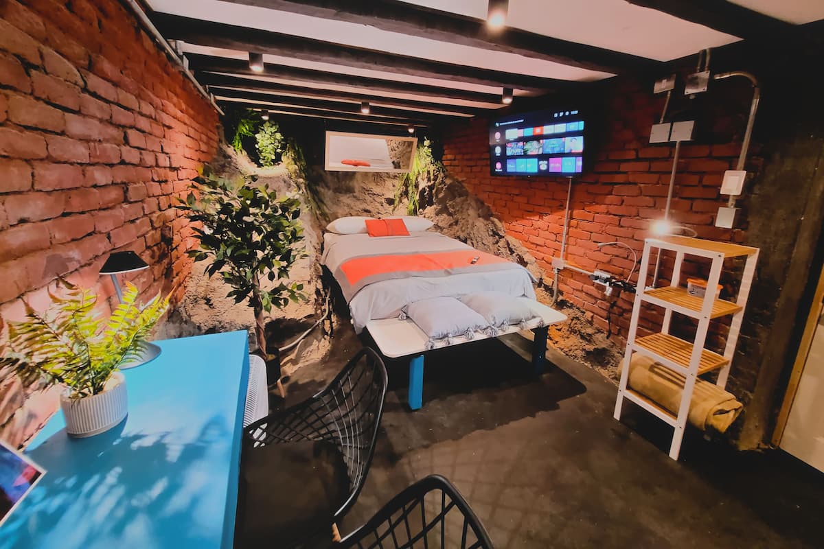 Quarto compacto com uma cama, escrivaninha, cadeira, televisão e prateleira.