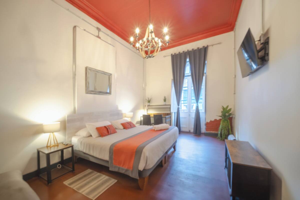 Quarto com cama, mesas de cabeceira, televisão, lustre e janela com cortinas.