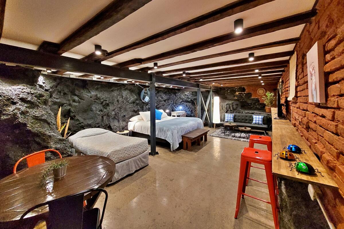 Quarto rústico com vigas aparentes, parede de pedra, cama, sofá, mesinha e uma cadeira.