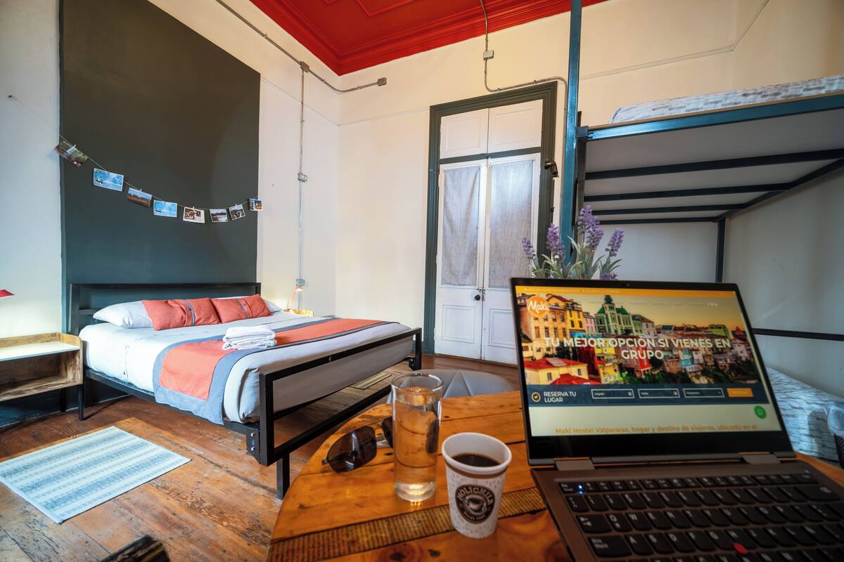 Um quarto com uma cama, um laptop sobre uma mesa, uma chávena e uma garrafa.
