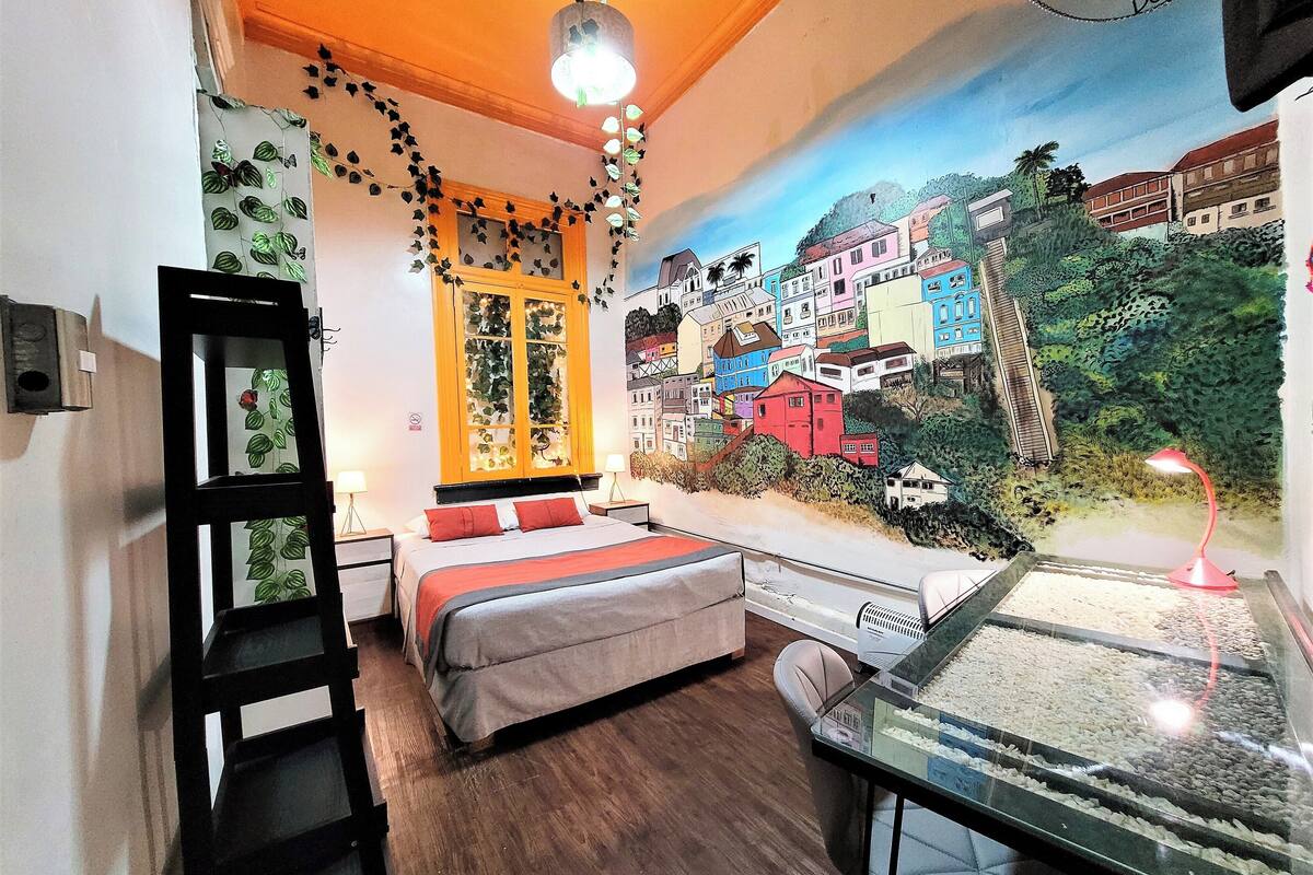 Quarto com cama, escrivaninha com aquário, janela com vista para edifícios coloridos e luminária no teto.