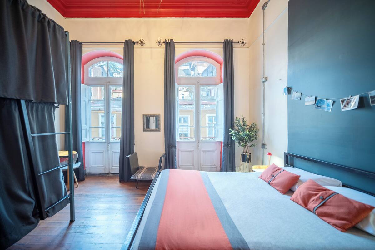 Quarto de hotel com uma cama, duas janelas com cortinas, uma cadeira, uma pequena mesa e uma parede com quadros.