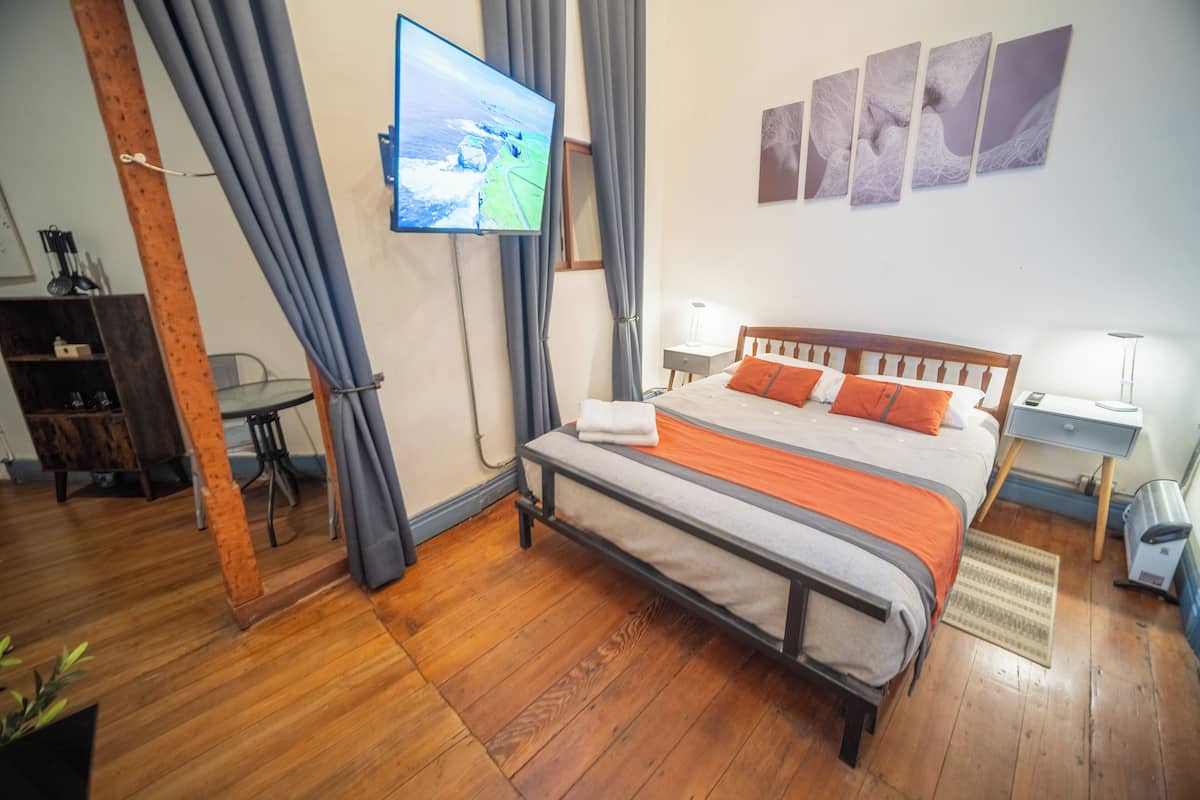 Quarto com cama de madeira, cortinas cinzas, televisão na parede e uma pequena escrivaninha com luminária.