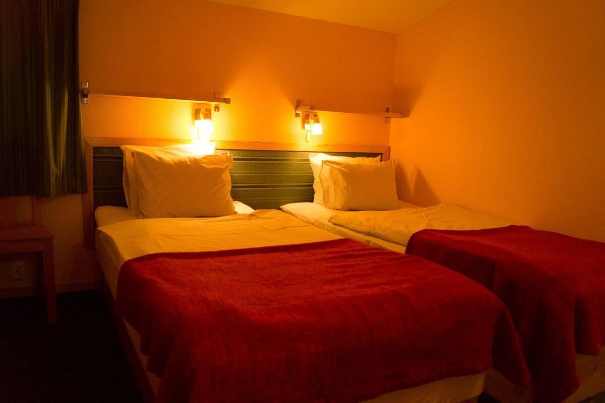 Habitación de hotel con dos camas, cada una con una manta roja, y dos lámparas fijadas a la pared.