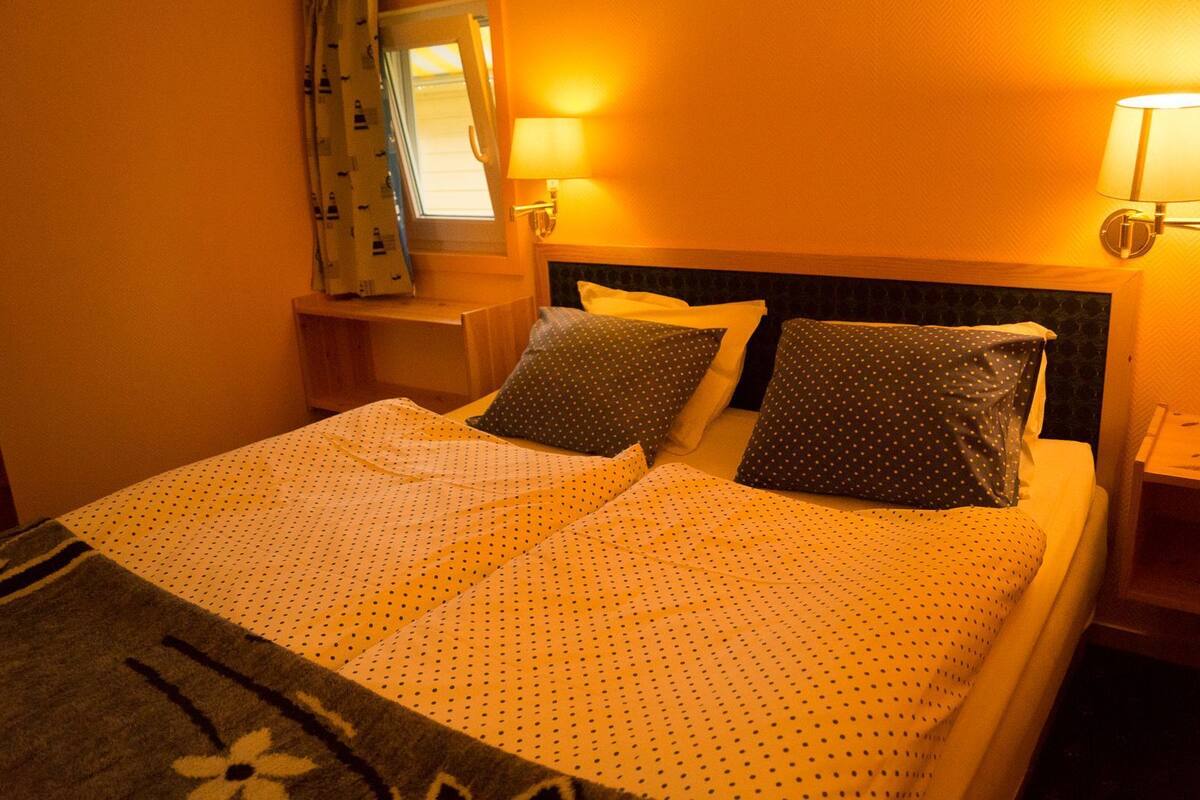 Habitación de hotel con cama, dos almohadas, mesita de noche, lámpara y ventana con cortinas.