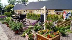 Il s’agit d’un bâtiment traditionnel, reconnaissable à une enseigne indiquant qu’il s’agit d’un hôtel. On y trouve un chariot en bois, ainsi qu’une variété de plantes et de fleurs en pot dans le jardin.