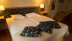 Un lit bien fait, avec du linge de lit blanc et deux coussins décoratifs, flanqué de tables de chevet avec des lampes, dans une pièce dont la tête de lit est ornée d’un motif floral et où l’on aperçoit un escalier au fond.