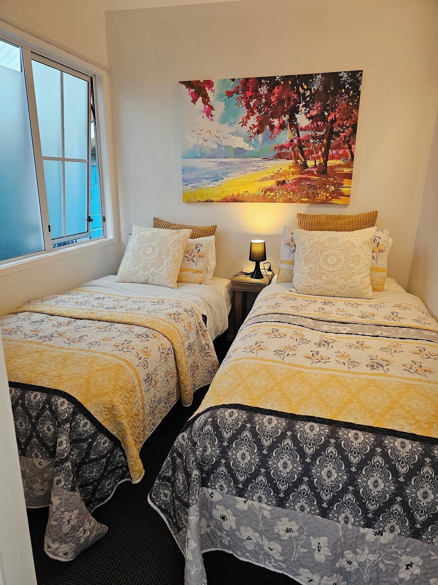 Quarto com duas camas, uma janela, um quadro e uma mesa de cabeceira com abajur.
