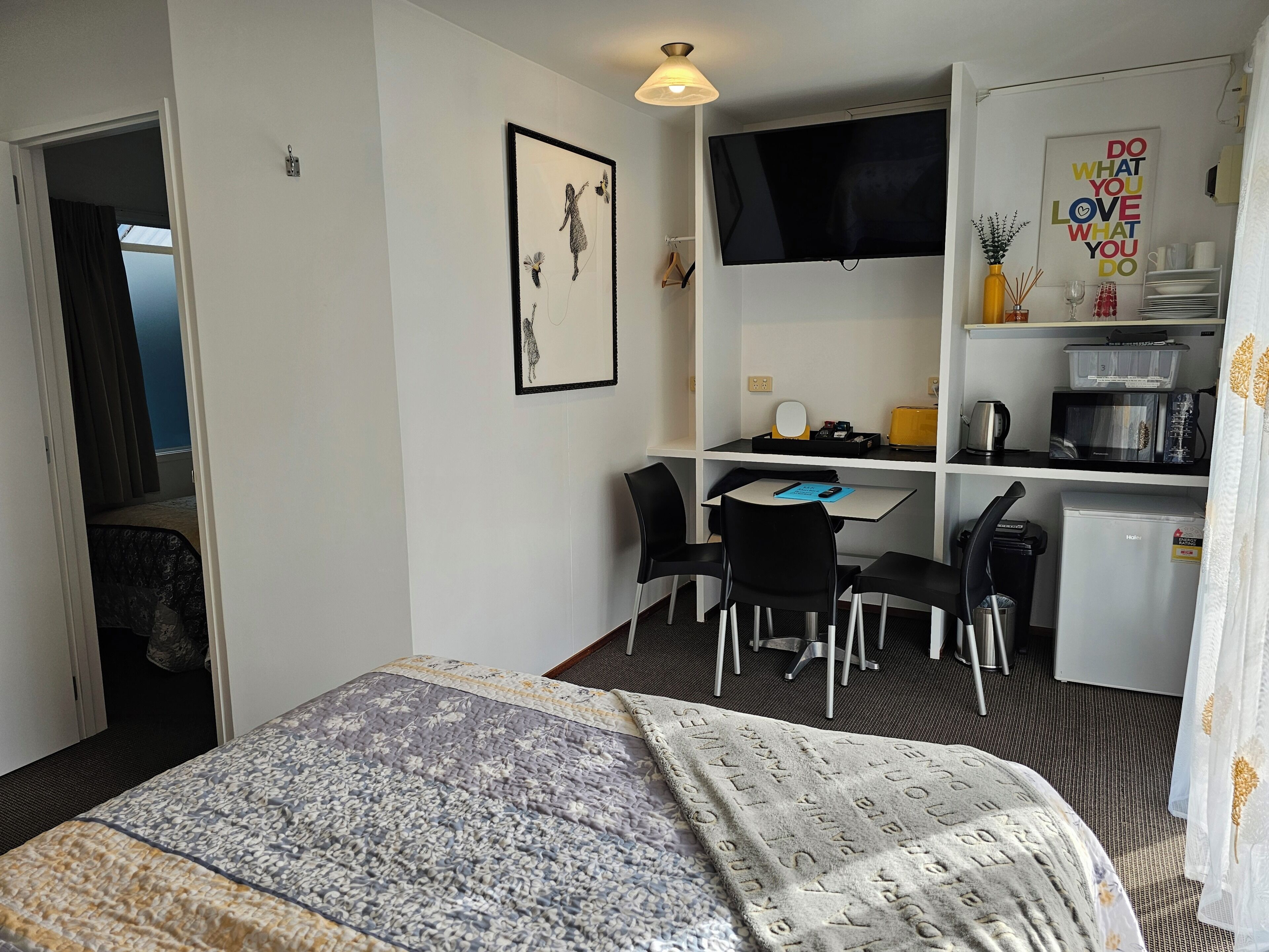 Habitación de hotel con cama, escritorio, sillas, televisor, kitchenette pequeña y puerta que conduce a otra habitación.