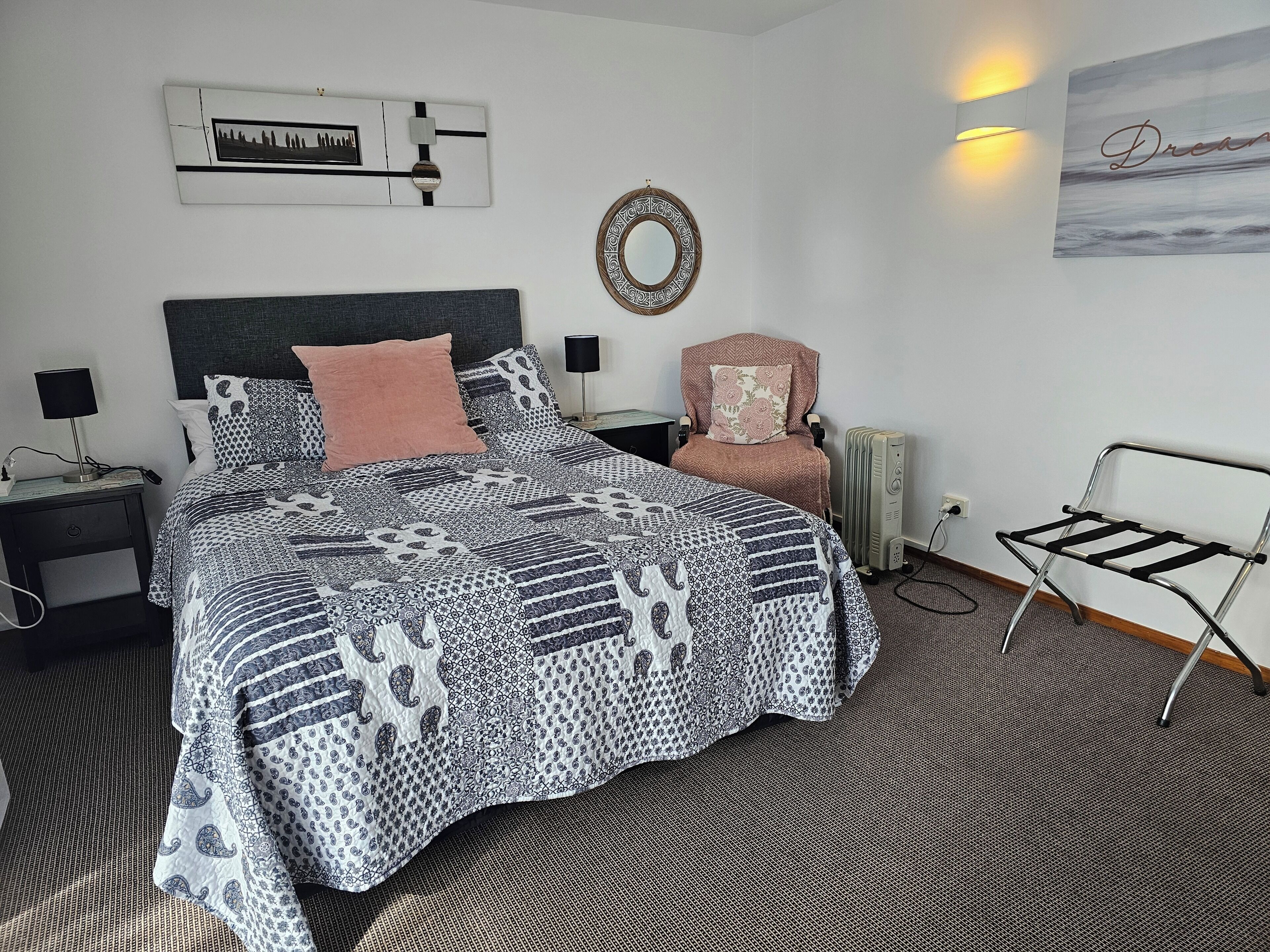 A neatly made bed with patterned bedding, a chair with a floral cushion, a wall clock, a mirror, and a radiator.