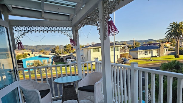 A covered outdoor area with a wooden deck, white wicker furniture, and a view of a residential neighborhood. - Albert Number 6 Motel (Whitianga)
