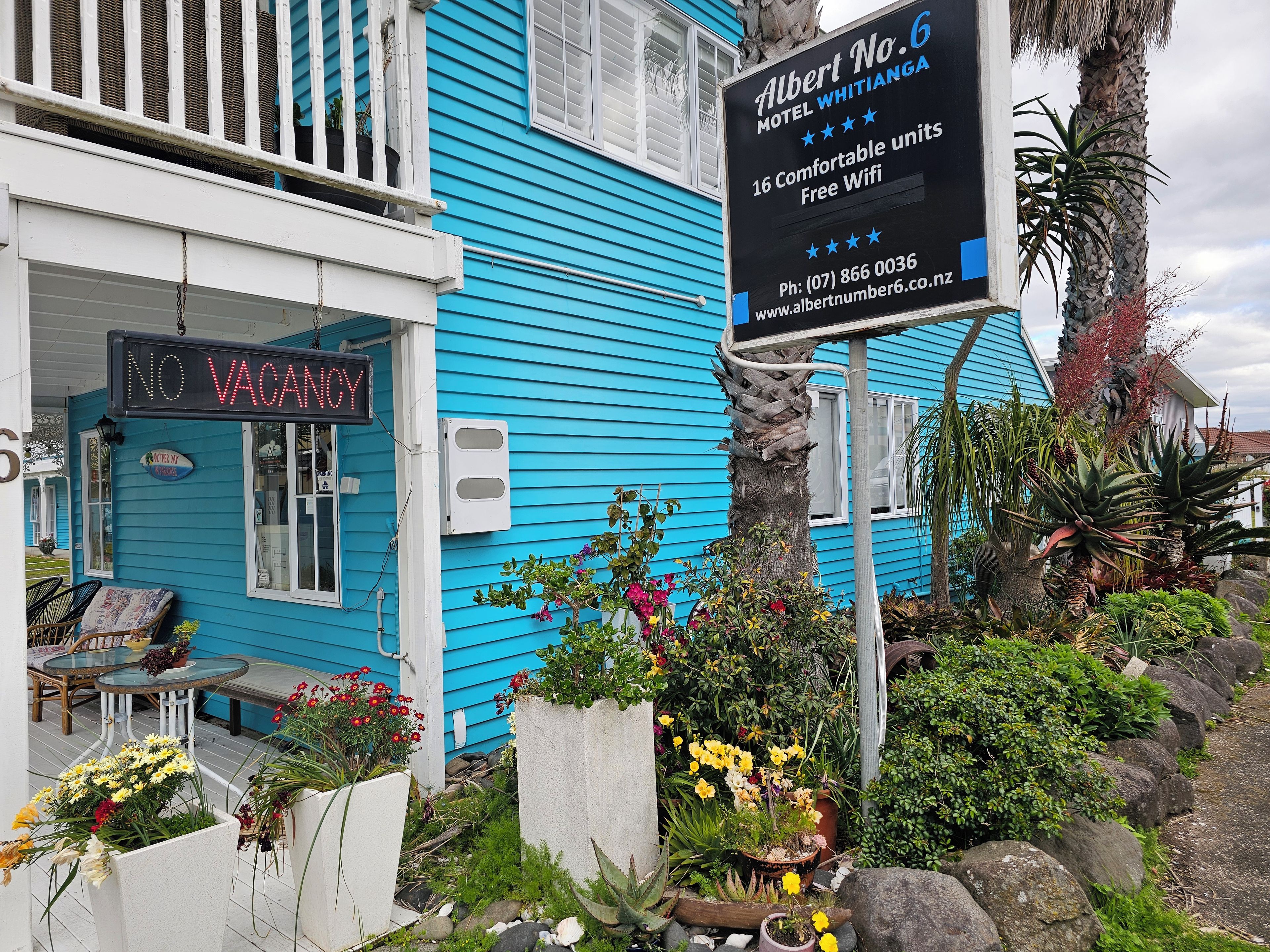 A motel with a neon 'NO VACANCY' sign, a signboard for Albert No.6 Motel Whitianga, and a variety of potted plants and flowers around the property.