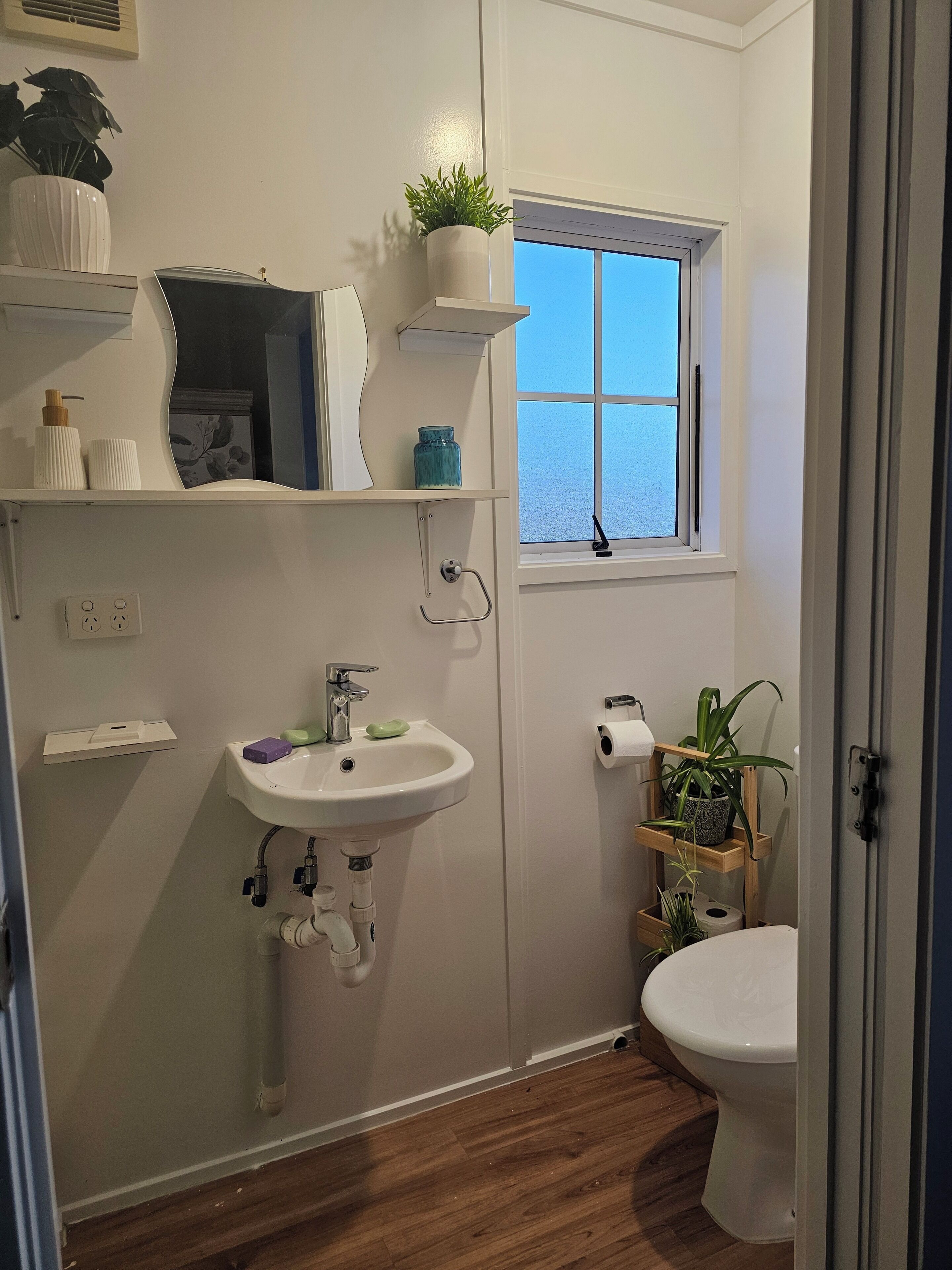 Un baño con lavamanos blanco, inodoro, ventana con vistas y estante con plantas.