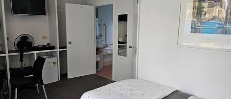 Un dormitorio con cama, escritorio con silla, televisor y baño visible a través de una puerta abierta.