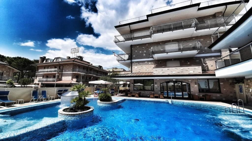 A swimming pool with a modern building in the background, a clear sky, and a few potted plants around the pool.