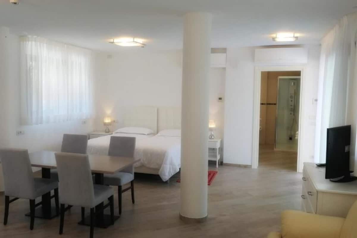 Une chambre d’hôtel moderne, équipée d’un lit, d’une table à manger avec des chaises, d’un téléviseur et d’une salle de bain visible à travers une porte ouverte.