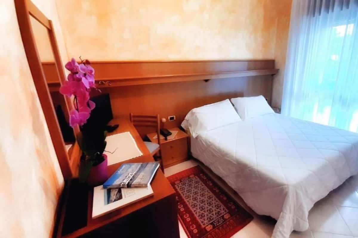 Une chambre d’hôtel comprenant un lit, un bureau avec une chaise, une petite table avec un vase de fleurs et une fenêtre avec des rideaux.