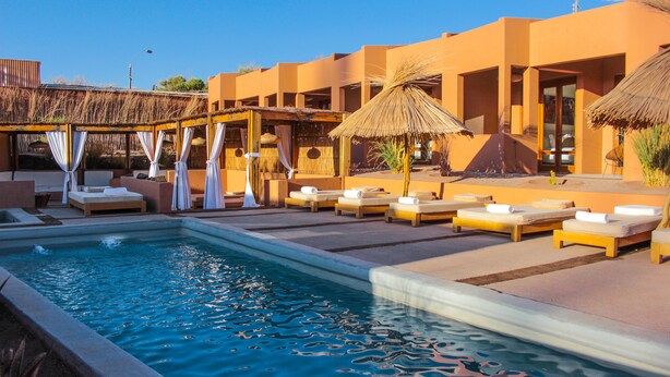 A pool area with lounge chairs and umbrellas, surrounded by a building with large windows and a thatched roof structure. - Noi Casa Atacama (San Pedro de Atacama)