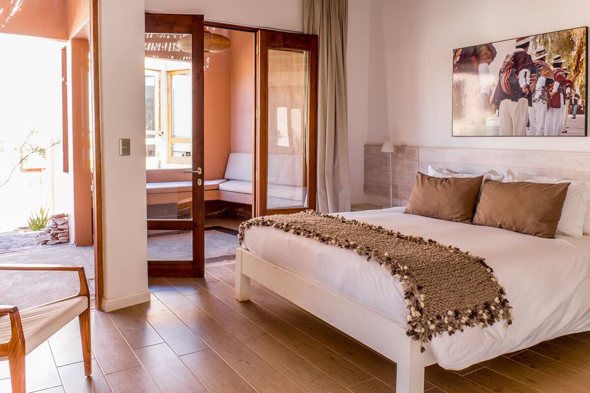 Een slaapkamer met een groot bed, een houten stoel, een deur die leidt naar een balkon en een schilderij aan de muur.
