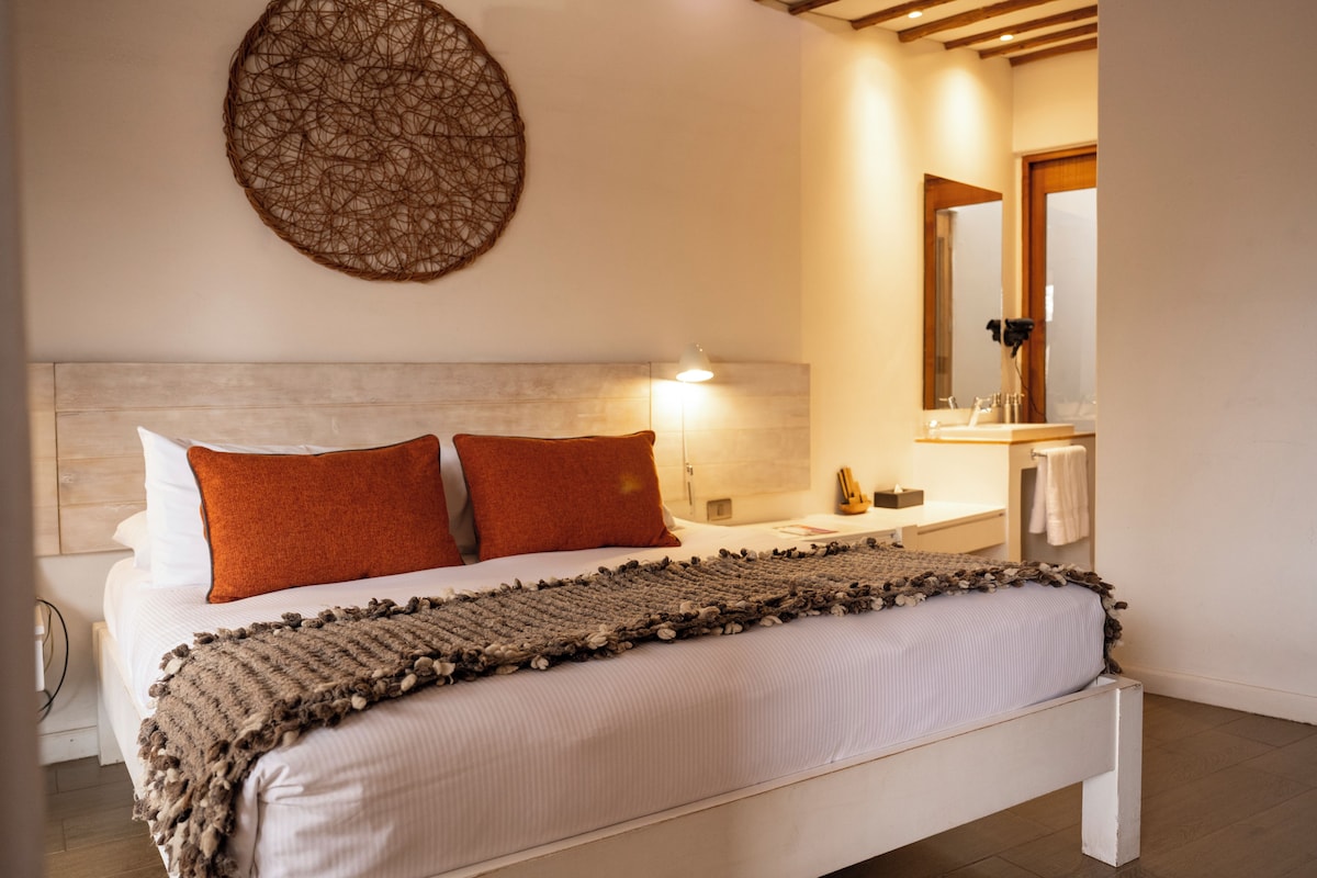 Een slaapkamer met een bed, kussens, een bedlampje, een spiegel en een klein nachtkastje.