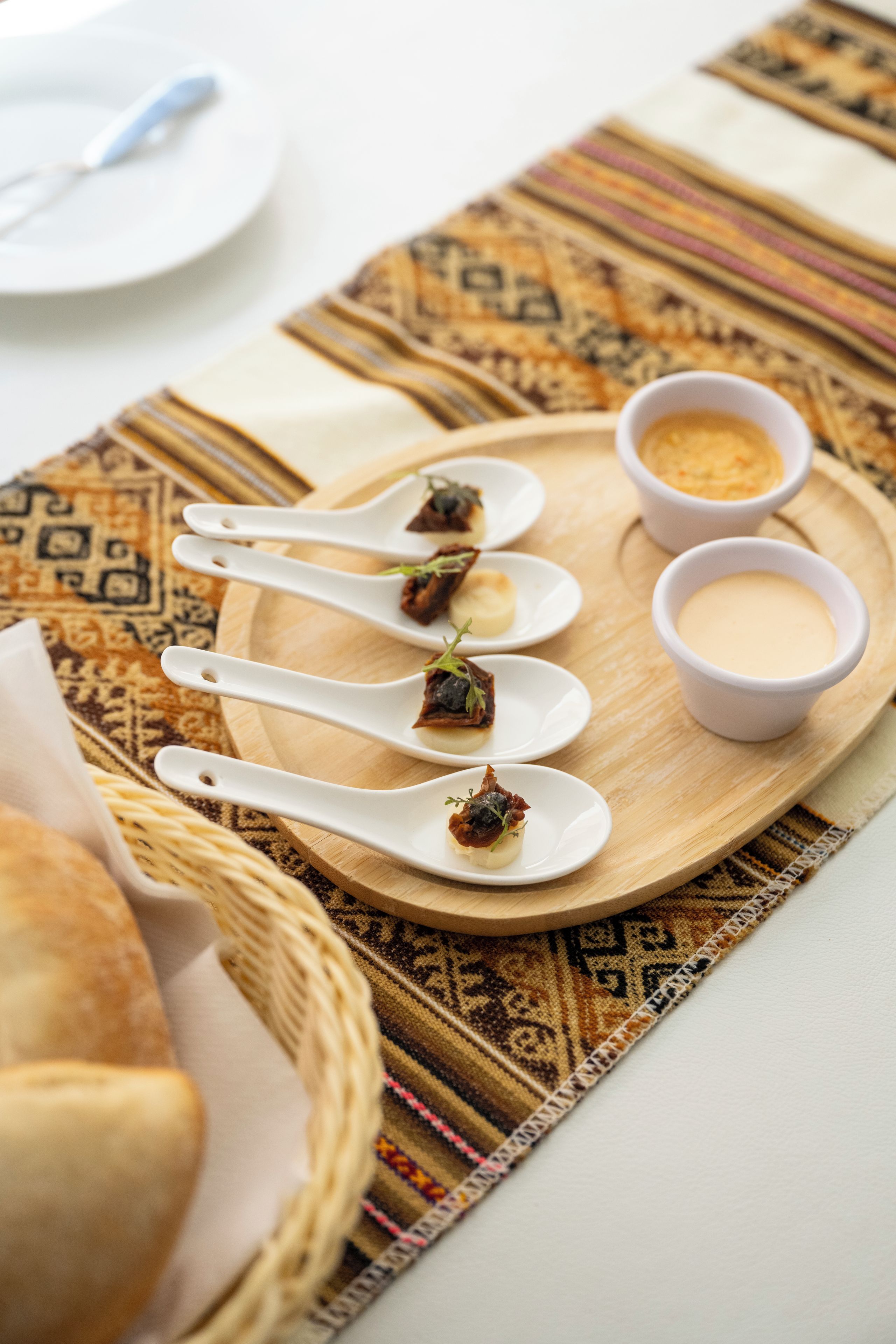 A wooden serving tray with white spoons and bowls containing food, placed on a patterned tablecloth.