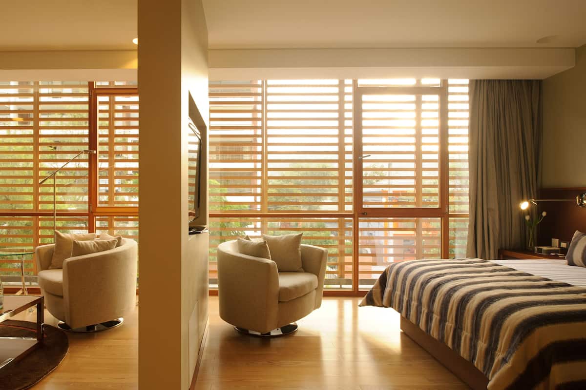 Une chambre d’hôtel avec un lit, deux fauteuils, un bureau et une grande fenêtre avec des stores.