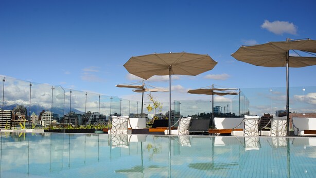Una piscina infinita en la azotea con sillones de descanso y sombrillas, con vistas a un paisaje urbano.