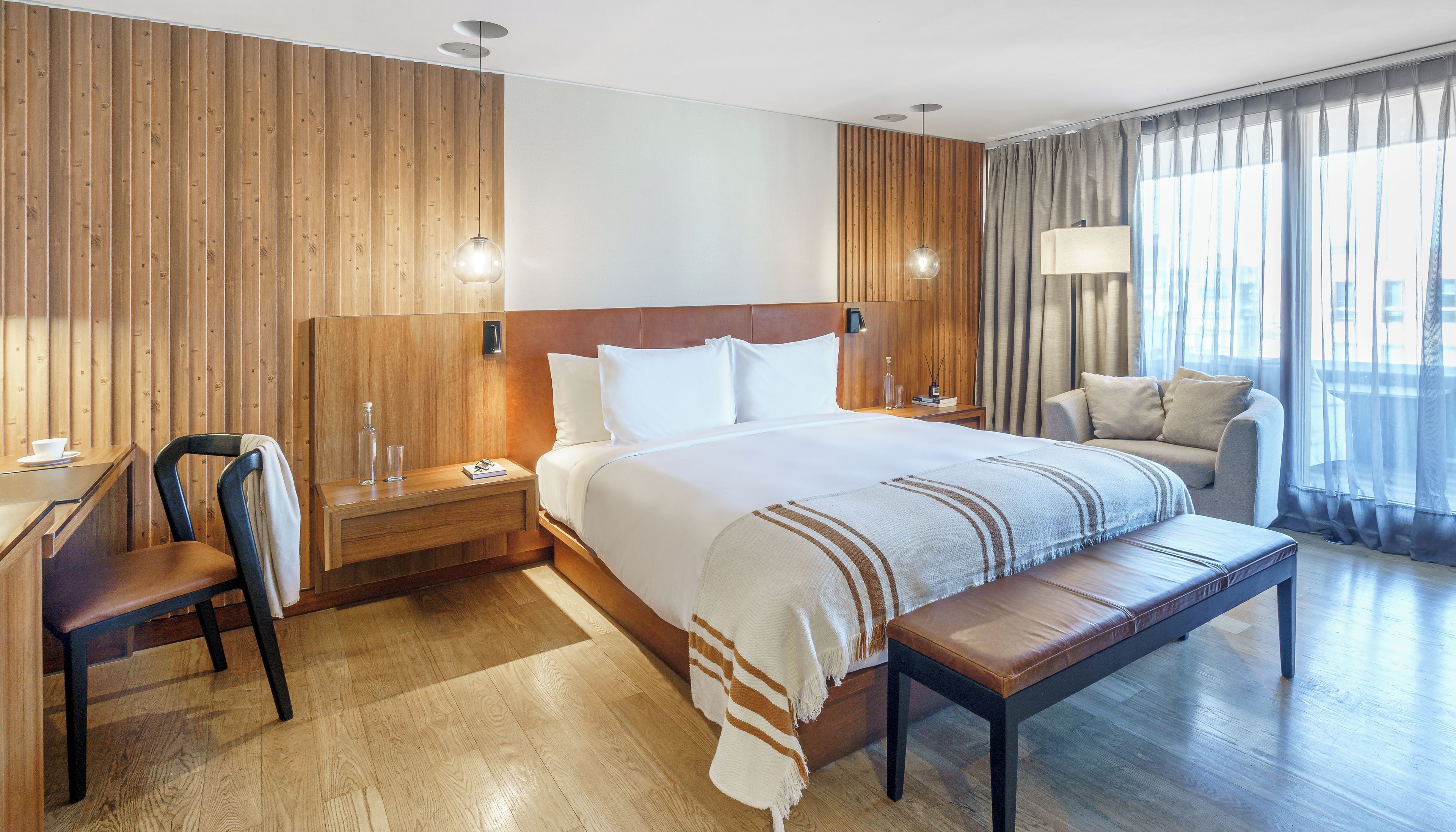 A hotel room with a wooden headboard, a large bed, a bench, a desk with a chair, and a sofa.