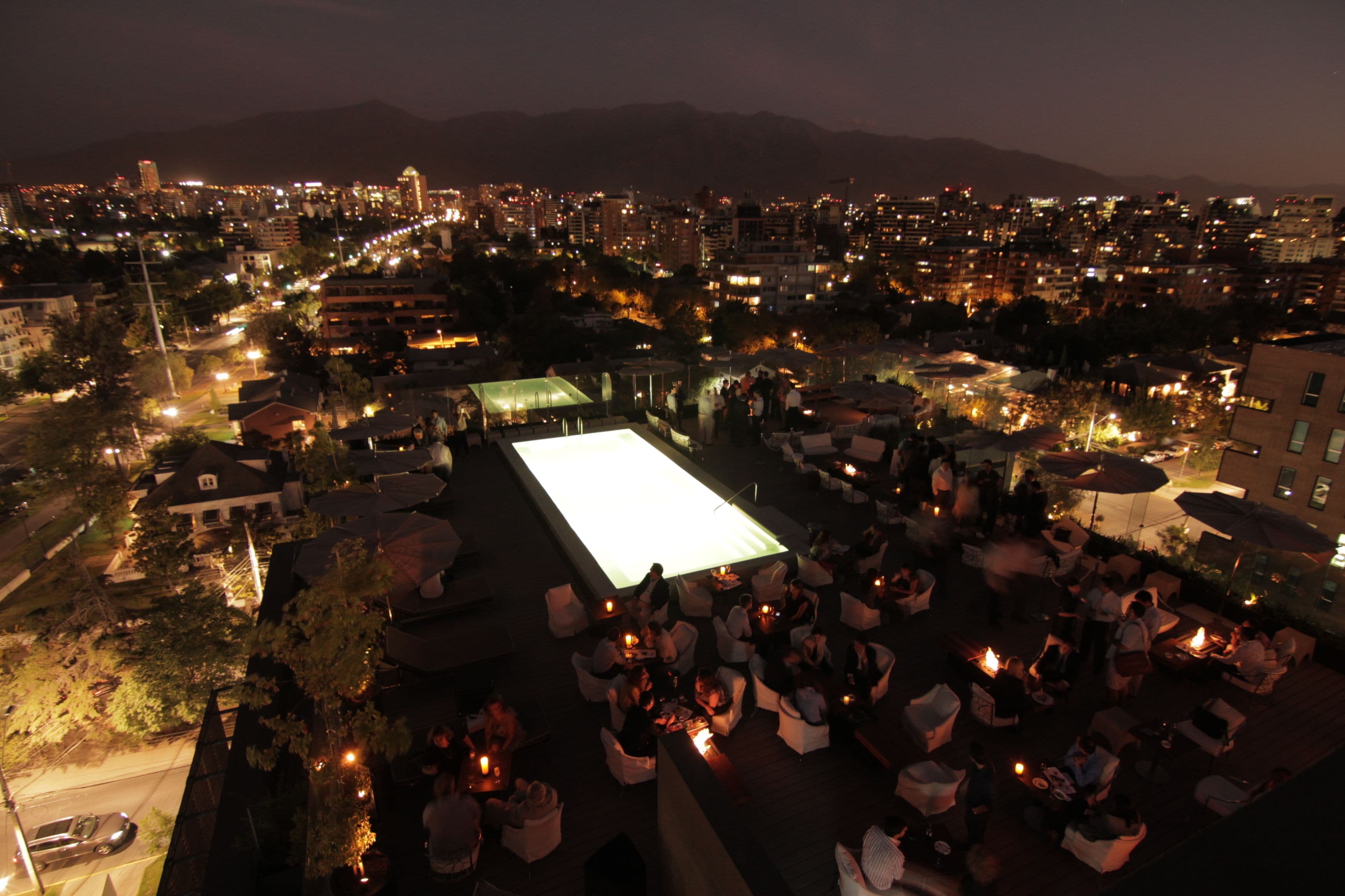 An outdoor rooftop bar with a large illuminated screen, seating areas, and a view of the cityscape at night.