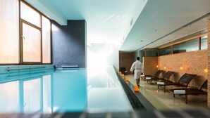 Una piscina interior moderna con pared de vidrio transparente, bancos de madera a un lado y una persona alejándose de la piscina.