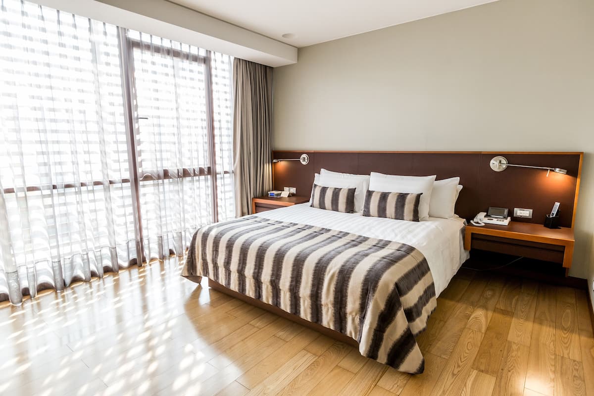 Une chambre d’hôtel avec un grand lit, un plancher en bois, une table de chevet avec une lampe et une fenêtre avec des rideaux.
