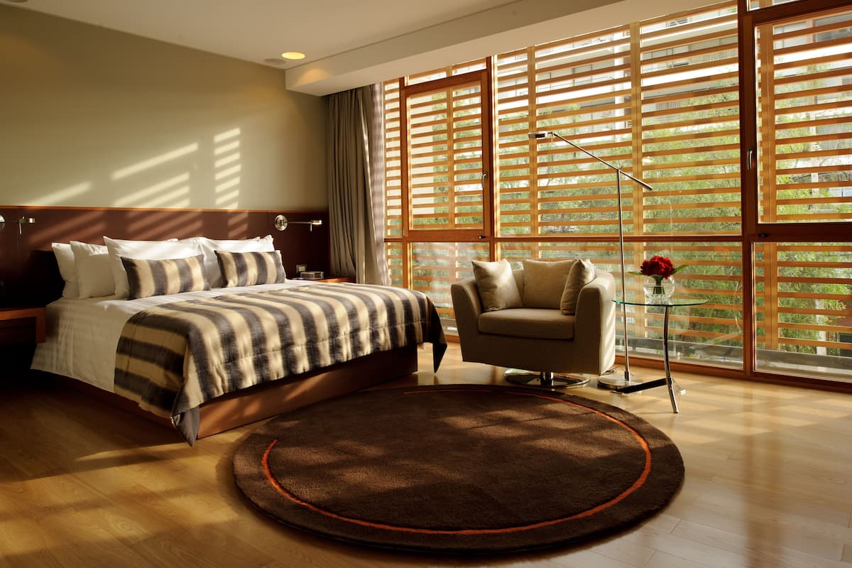 Une chambre moderne avec un grand lit, un coin salon avec un fauteuil et une grande fenêtre équipée de stores.