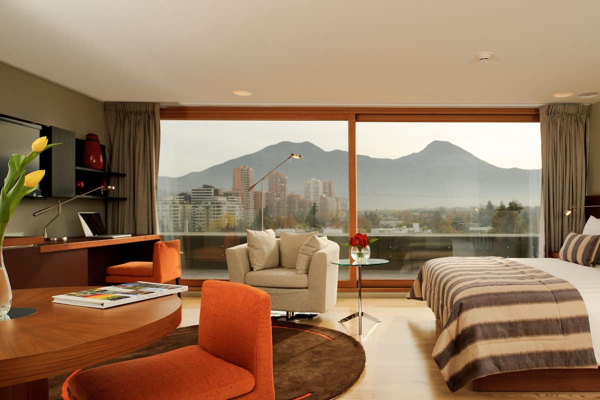 Une chambre d’hôtel équipée d’un lit, d’un bureau, d’une chaise, d’une télévision et offrant une vue sur un paysage urbain avec des montagnes.
