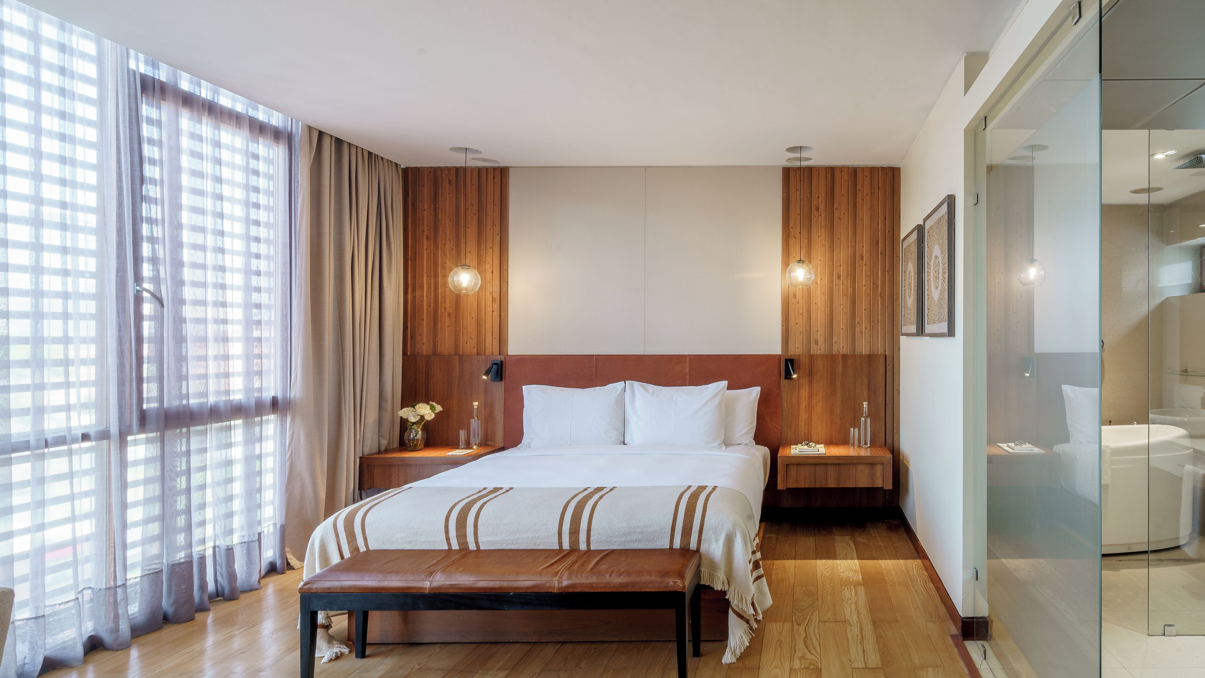 Een hotelkamer met een bed, nachtkastjes, een bankje, een groot raam met gordijnen, en een zichtbare badkamer achter een glazen wand.