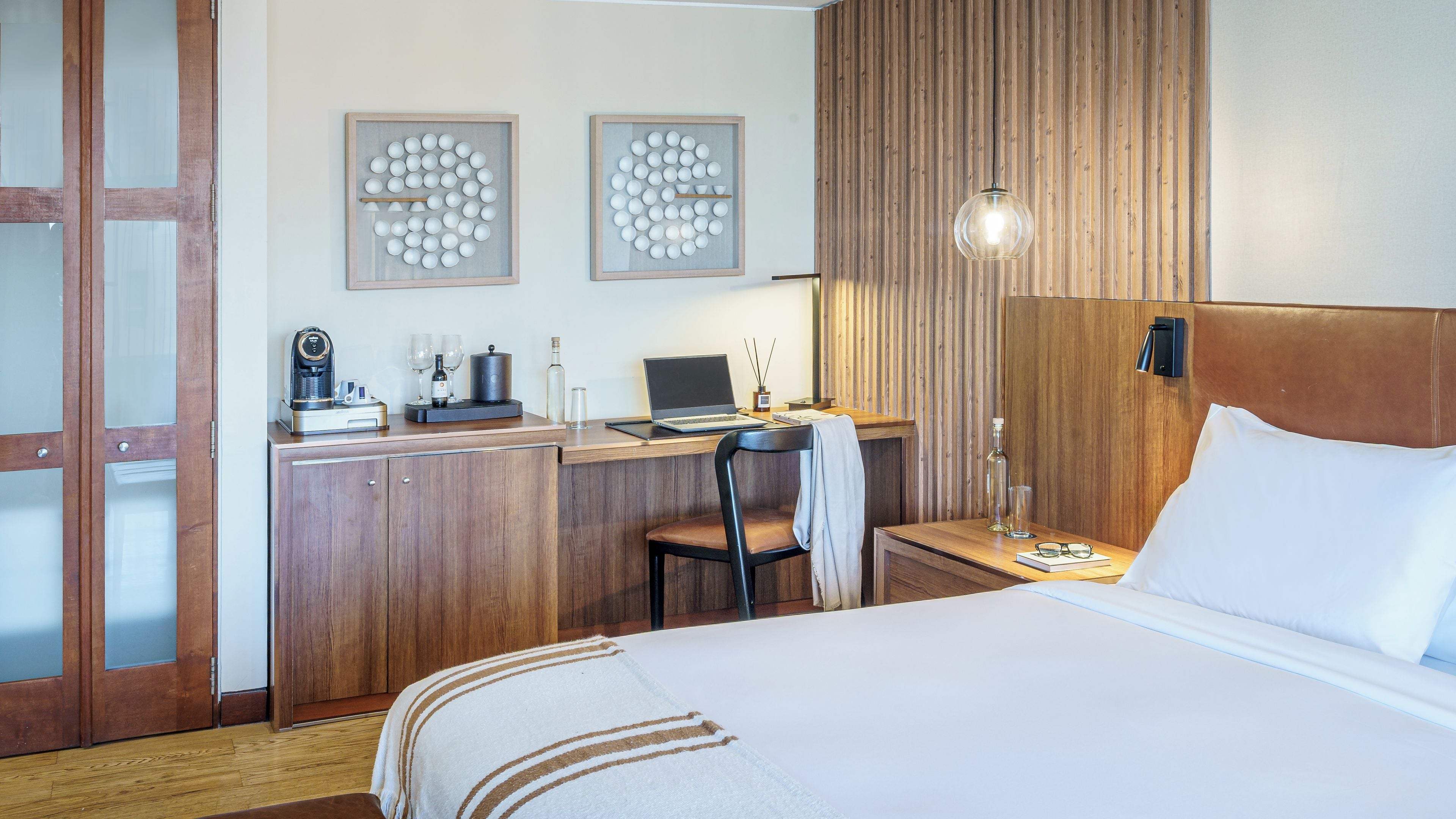 A hotel room with a bed, a desk with a laptop, a coffee maker, and a chair. There are two framed wall art pieces and a wooden panel wall.