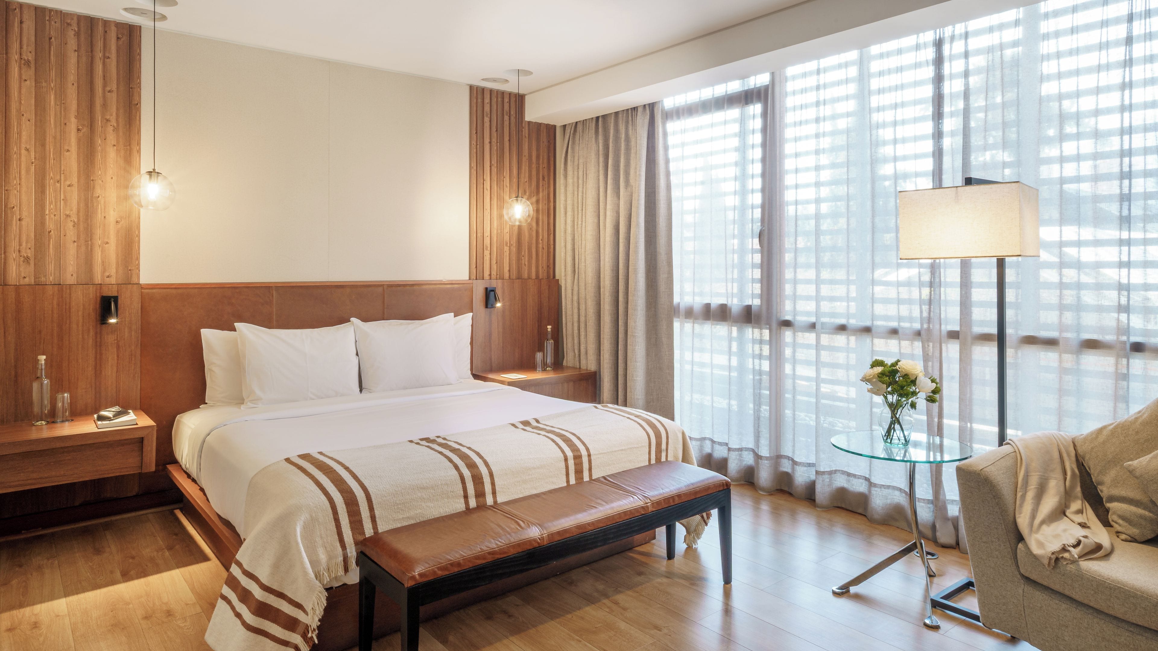 Habitación de hotel con una cama grande, cabecera de madera, mesitas de noche, un banco, una mesa de vidrio con una lámpara y vistas a través de cortinas translúcidas.