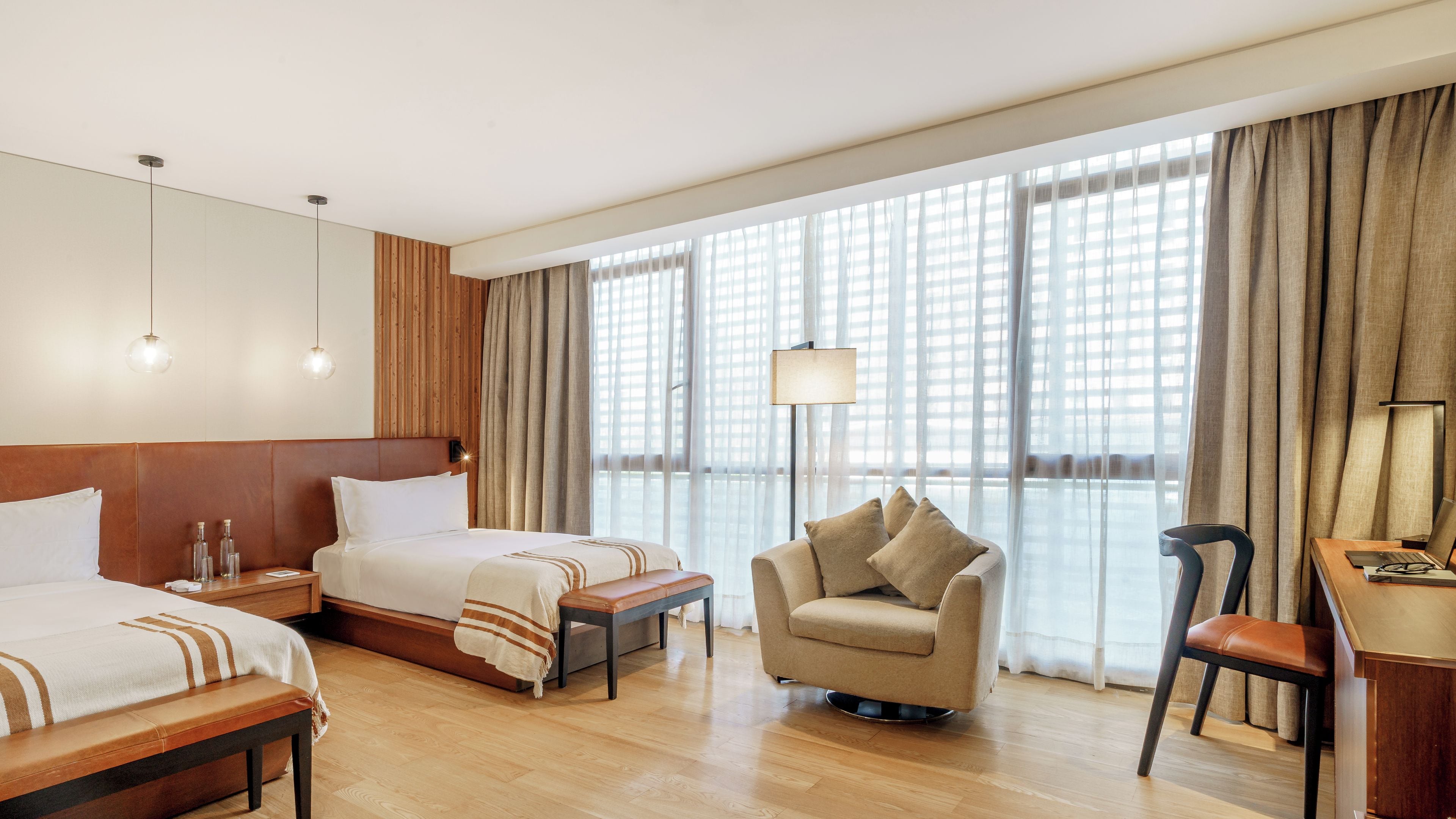 Habitación de hotel con dos camas, un sofá, un escritorio con silla y ventanas amplias con cortinas.