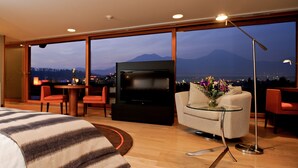 A modern hotel room with a large bed, a TV, a glass table, and a view of mountains and city lights. - Noi Vitacura (Santiago)