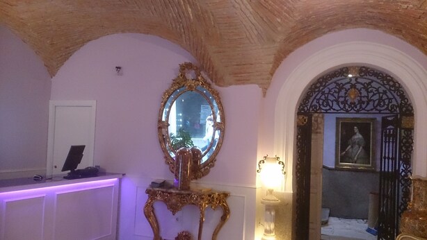 Zona de recepción con un mostrador iluminado en morado, un espejo grande y ornamentado, y un retrato en un arco.