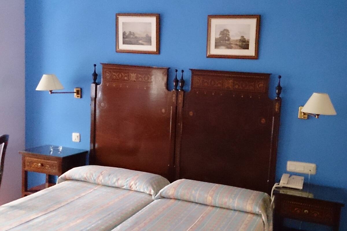 A room with a bed, two wall-mounted lamps, two framed pictures, and a bedside table with a phone.