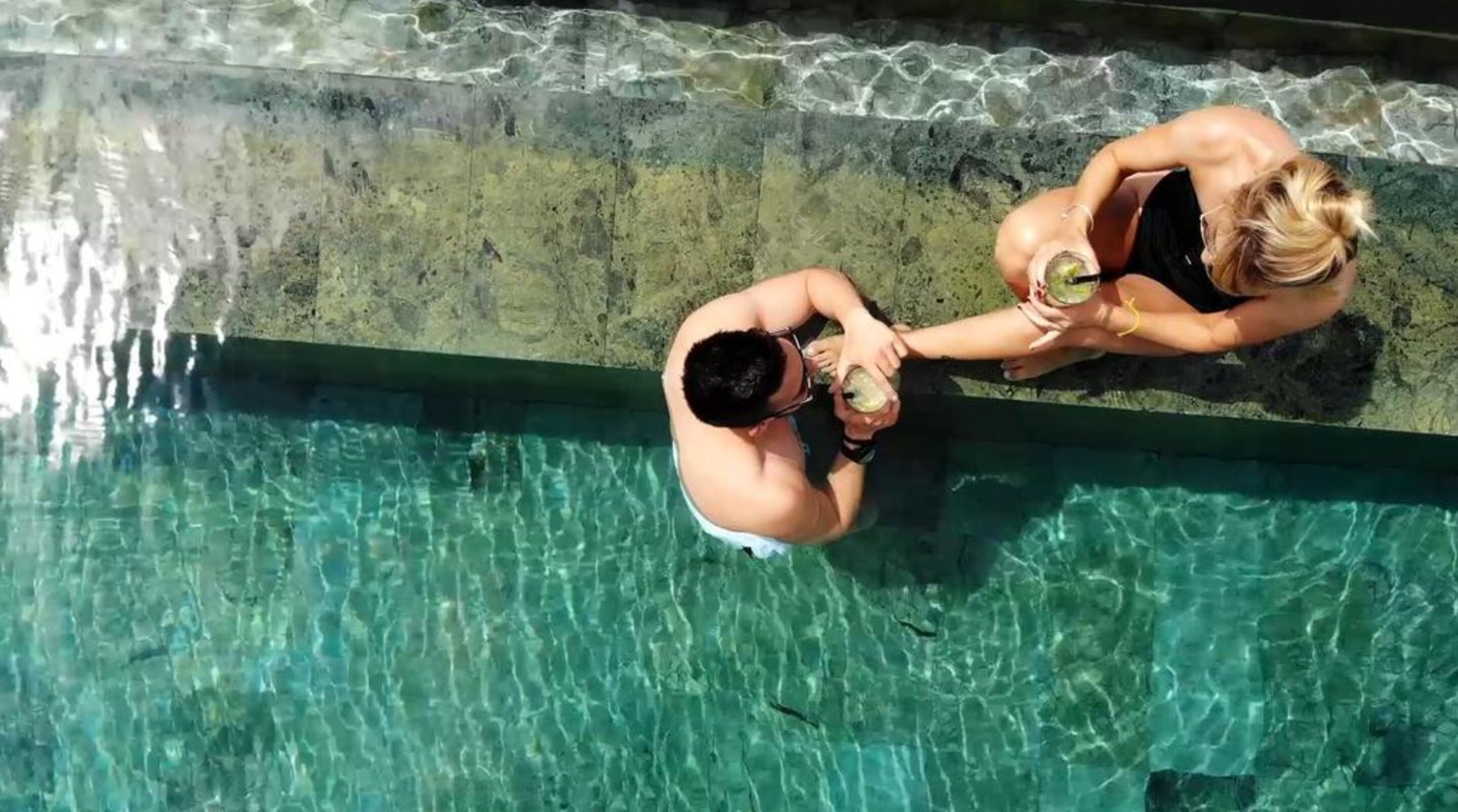 A clear view of a swimming pool with two people sitting on the edge, one holding a drink.