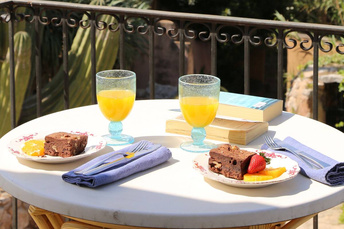 Uma mesa com um livro, dois copos de suco de laranja, um prato com um brownie e morangos, e uma toalha dobrada numa varanda com vista para um jardim com cactos e palmeiras.