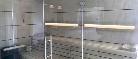 A modern, glass-walled sauna with a bench, a small heater, and a clock on the wall.